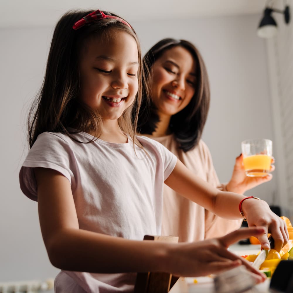 A woman holding a glass of orange juice is standing behind a young girl as she is preparing food.