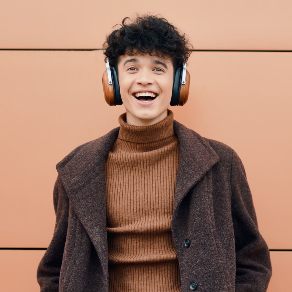 A young man is standing outside wearing headphones and smiling really big.