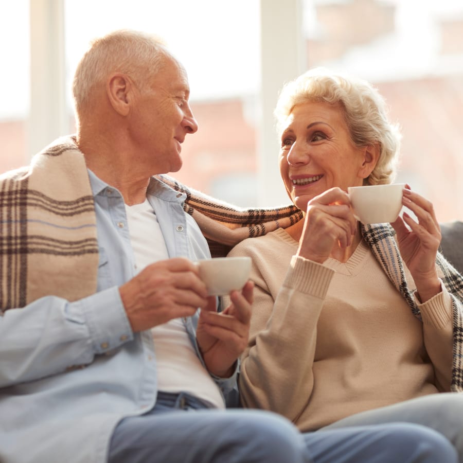 An older man and woman are sitting on a sofa with a branket around them drinking a warm beverage.