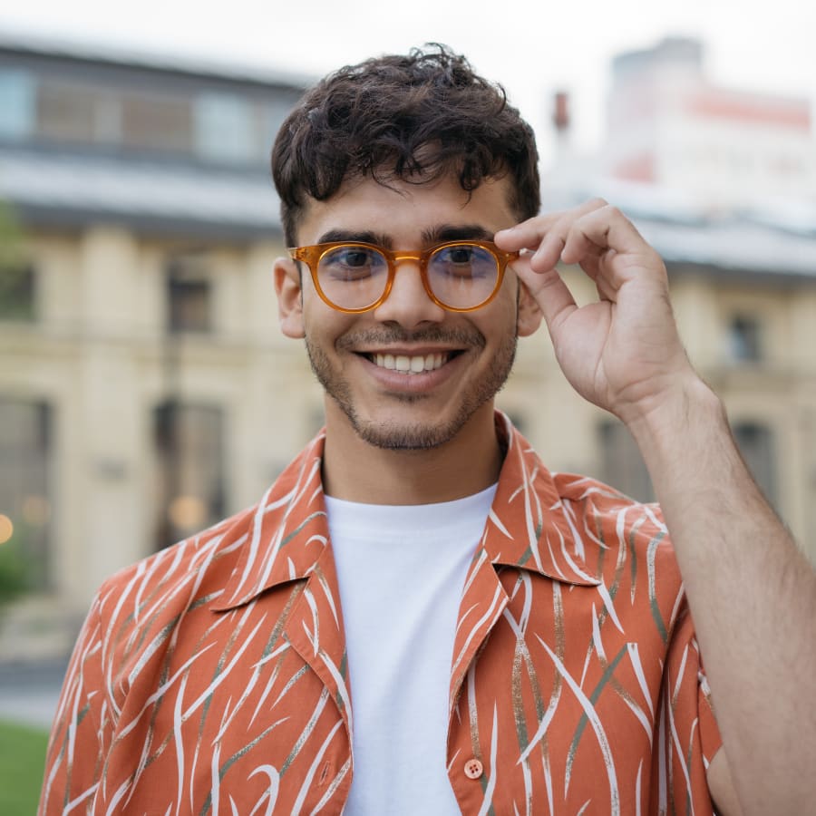 A young man is standing outside smiling and holding the rim of his glasses with his hand..