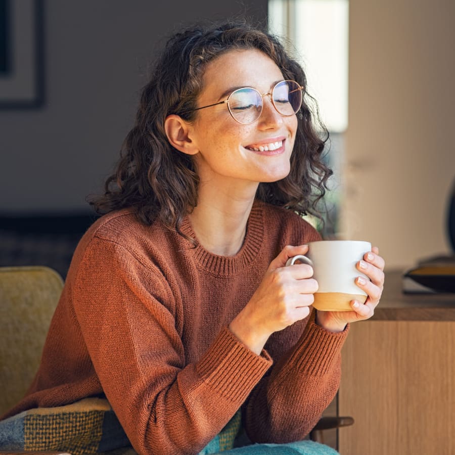 A young woman with glasses is sitting and smiling while holding a cofee mug in her hands.