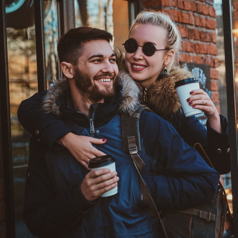 A woman is embracinga man from behind. Both are dressed warmly and holding take out coffee cups.
