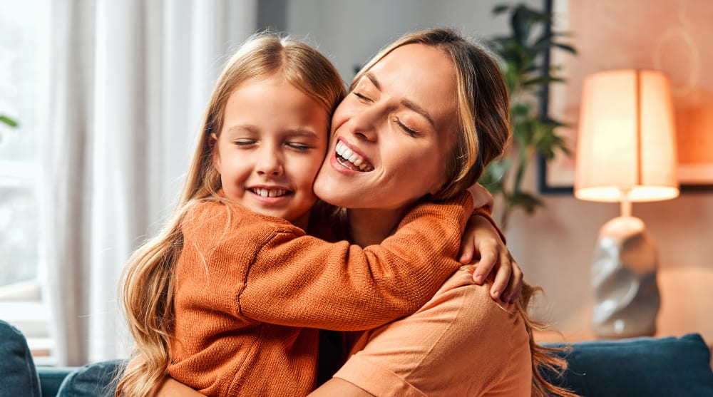 A woman hugs a young girl tightly while sitting inside a home.